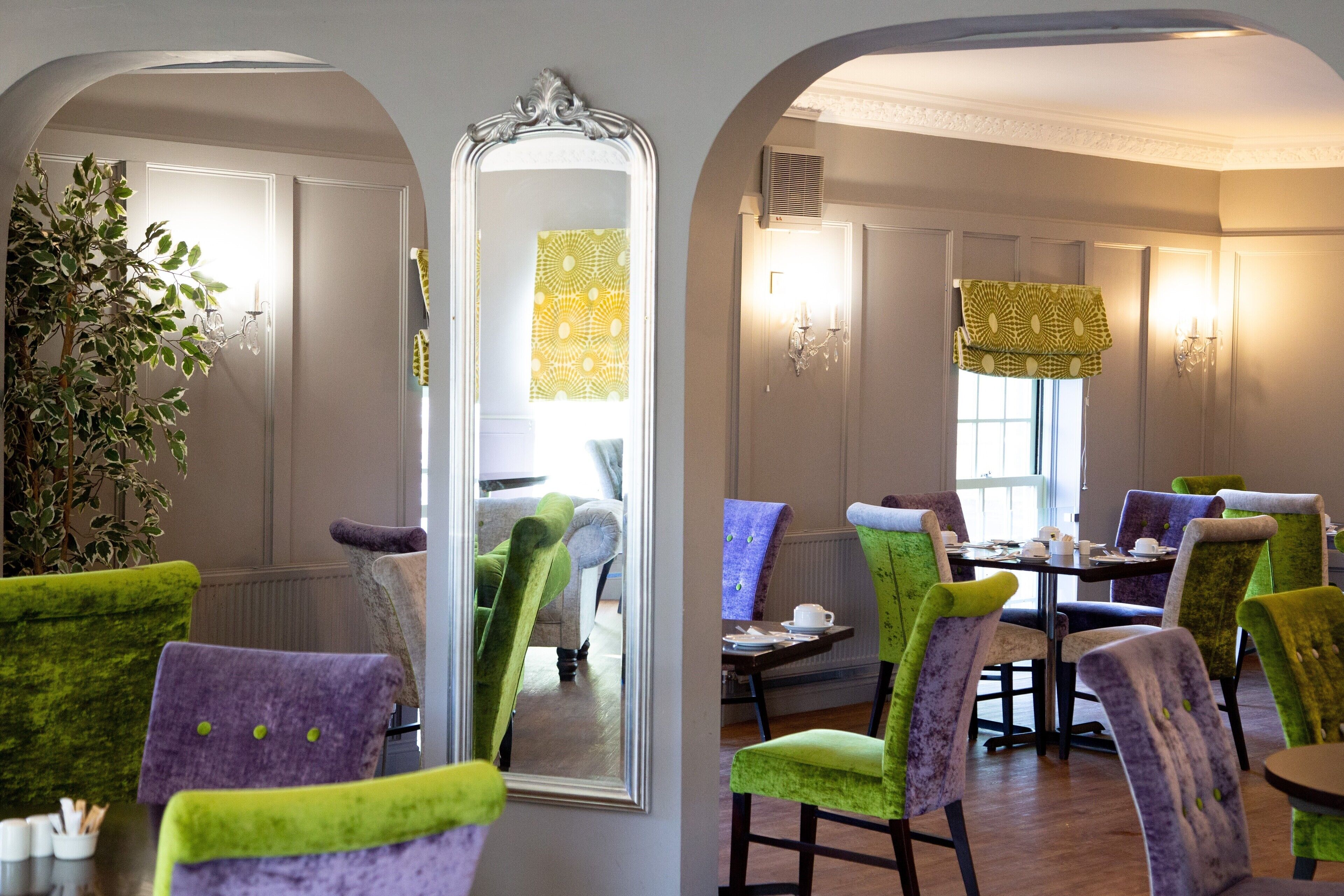A dining area with a table set for four, surrounded by colorful chairs, a large mirror, and a potted plant.