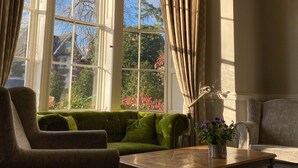 Ein Zimmer mit einem grünen Sofa, einem Holztisch und einem Stuhl. Große Fenster mit Vorhängen sind vorhanden.