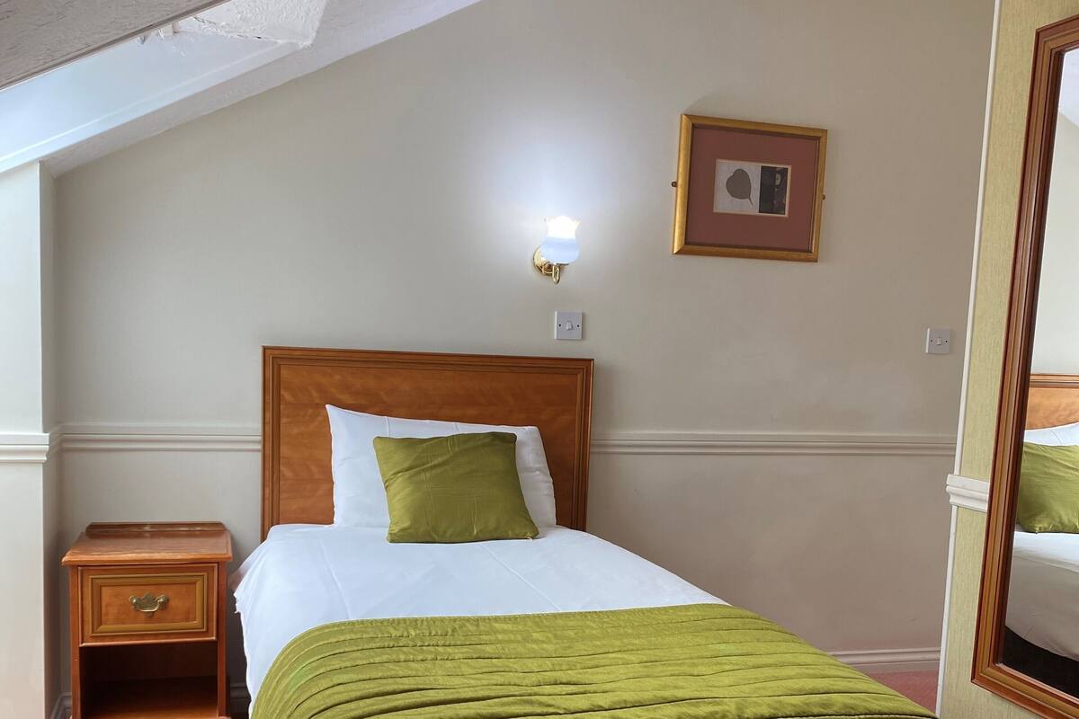 A neatly made bed with a green blanket, a wooden nightstand with a drawer, a wall-mounted light, and a framed picture.