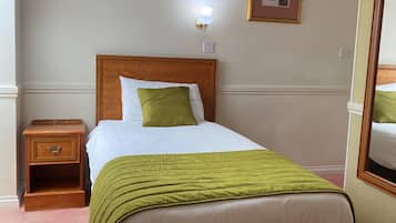 A neatly made bed with a green blanket, a wooden nightstand with a drawer, a wall-mounted light, and a framed picture.