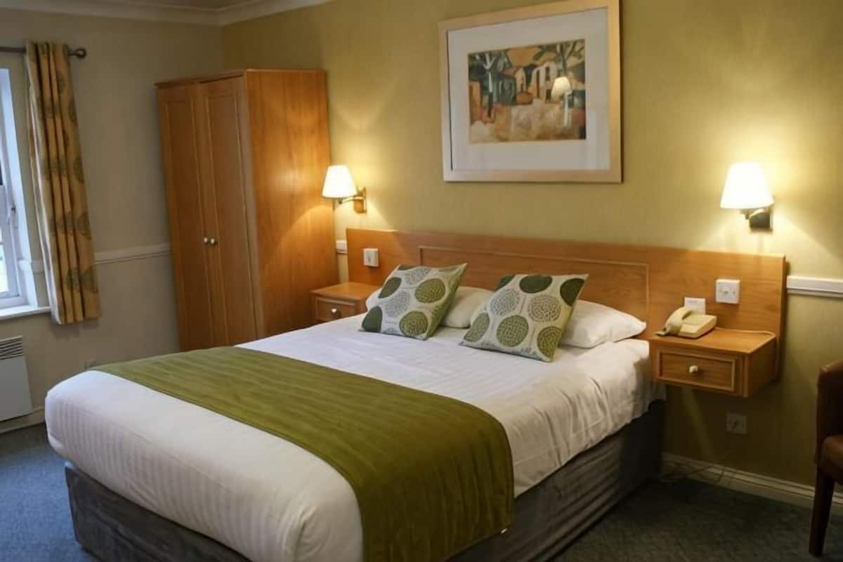 A hotel room with a bed, two bedside lamps, a nightstand, a telephone, and a painting on the wall.