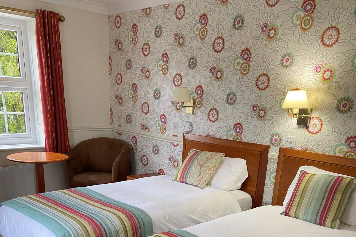 A hotel room with two single beds, a wooden headboard, a small round table, a chair, and a floral wallpaper.