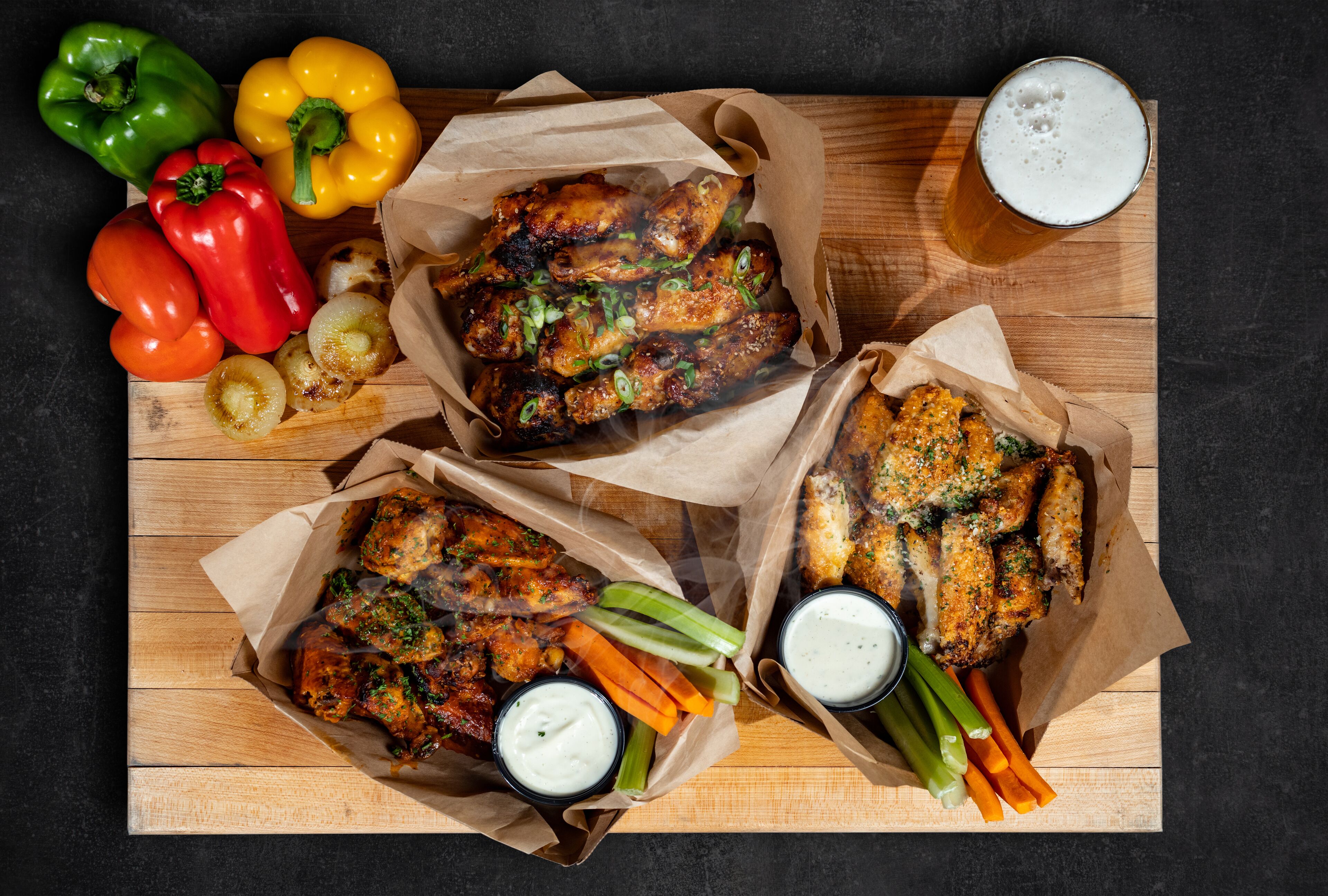 Uma mesa de madeira com três cestos de papel recheados com asas de frango, aipo, cenouras e dois molhos para acompanhar.