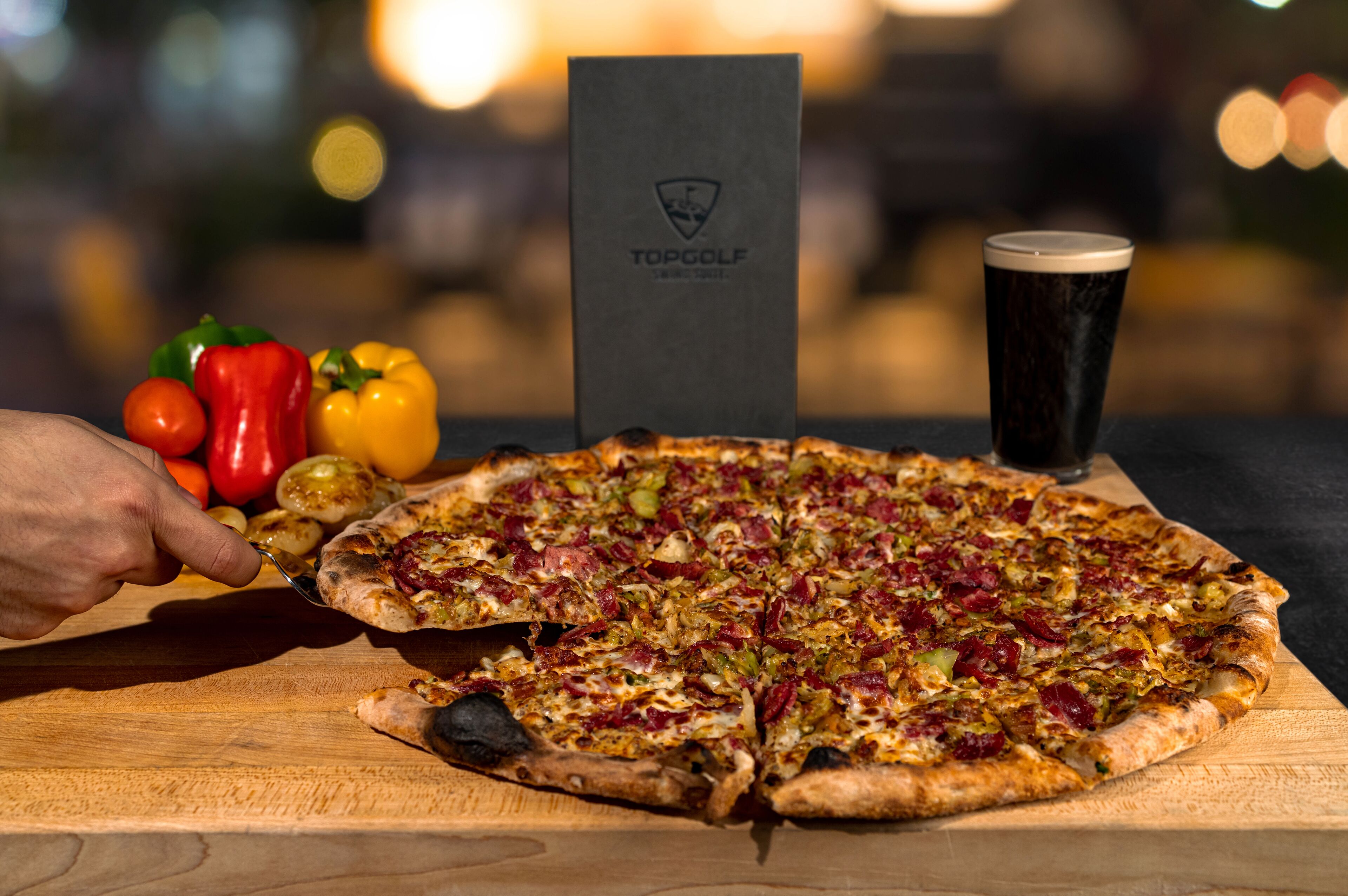 Pizza op een houten plank, met een drankje en groenten op de achtergrond.