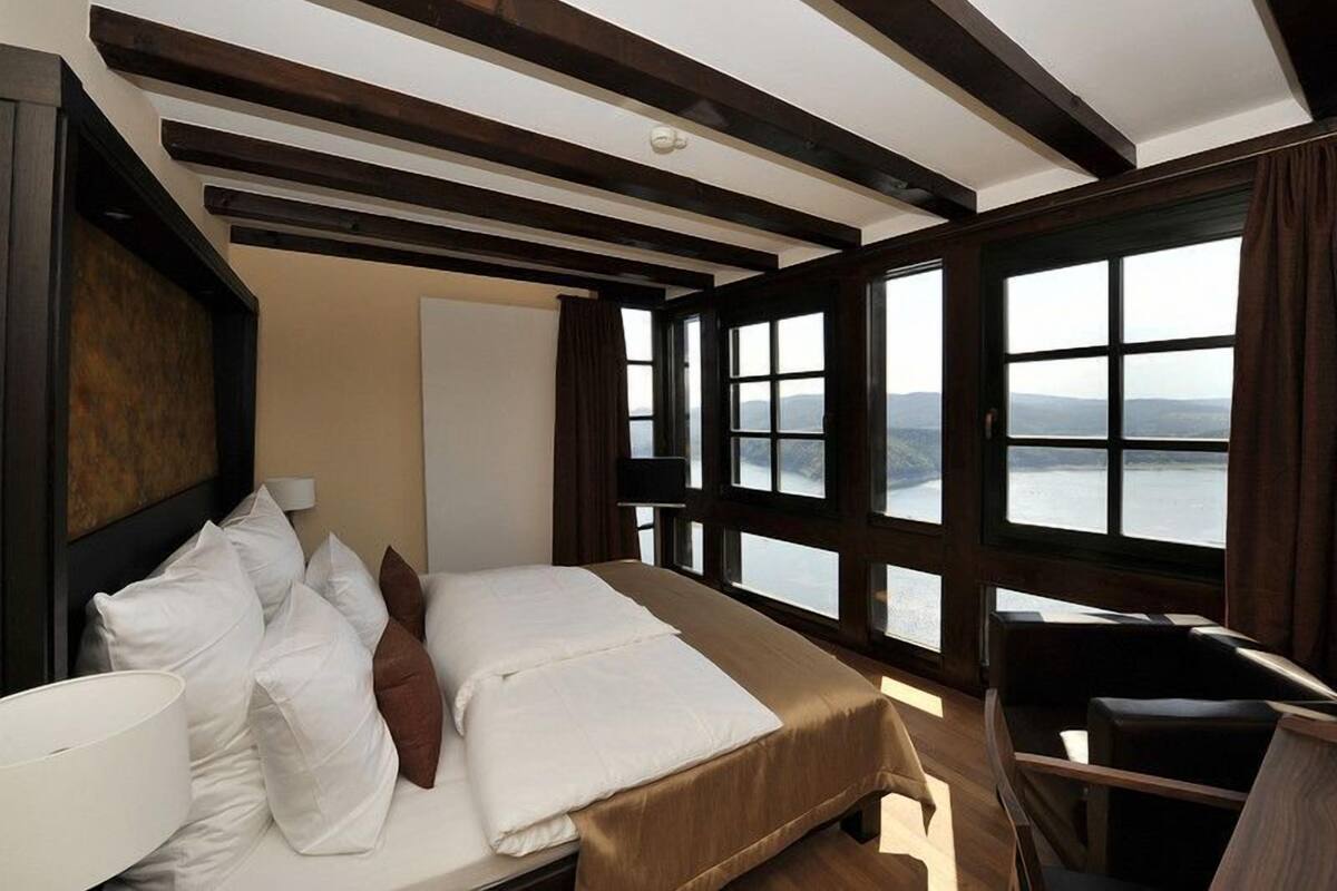 A bedroom with a bed, a desk, and large windows offering a view of the outdoors.