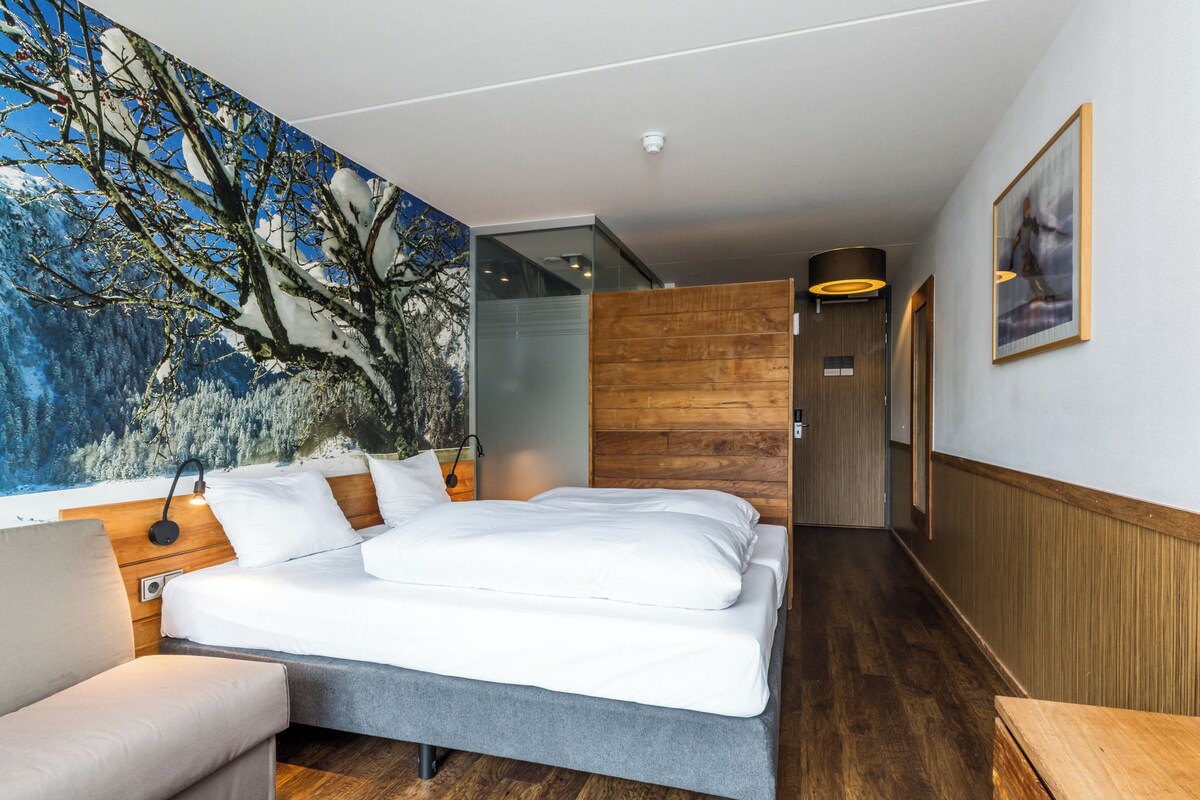 A modern hotel room with a large bed, a sofa, a wooden accent wall, and a mural of trees.