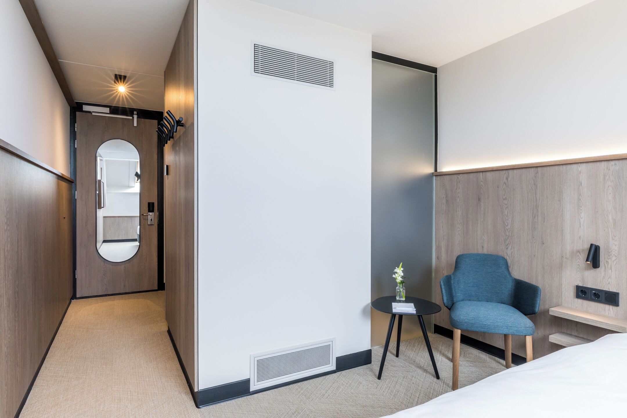 Une chambre d’hôtel moderne comprenant un lit, une petite table avec un vase, une chaise, un miroir et un luminaire fixé au mur.