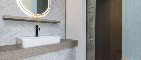 Un baño moderno con un lavamanos rectangular, un espejo con un estante de madera encima y una zona de ducha con puerta de vidrio.