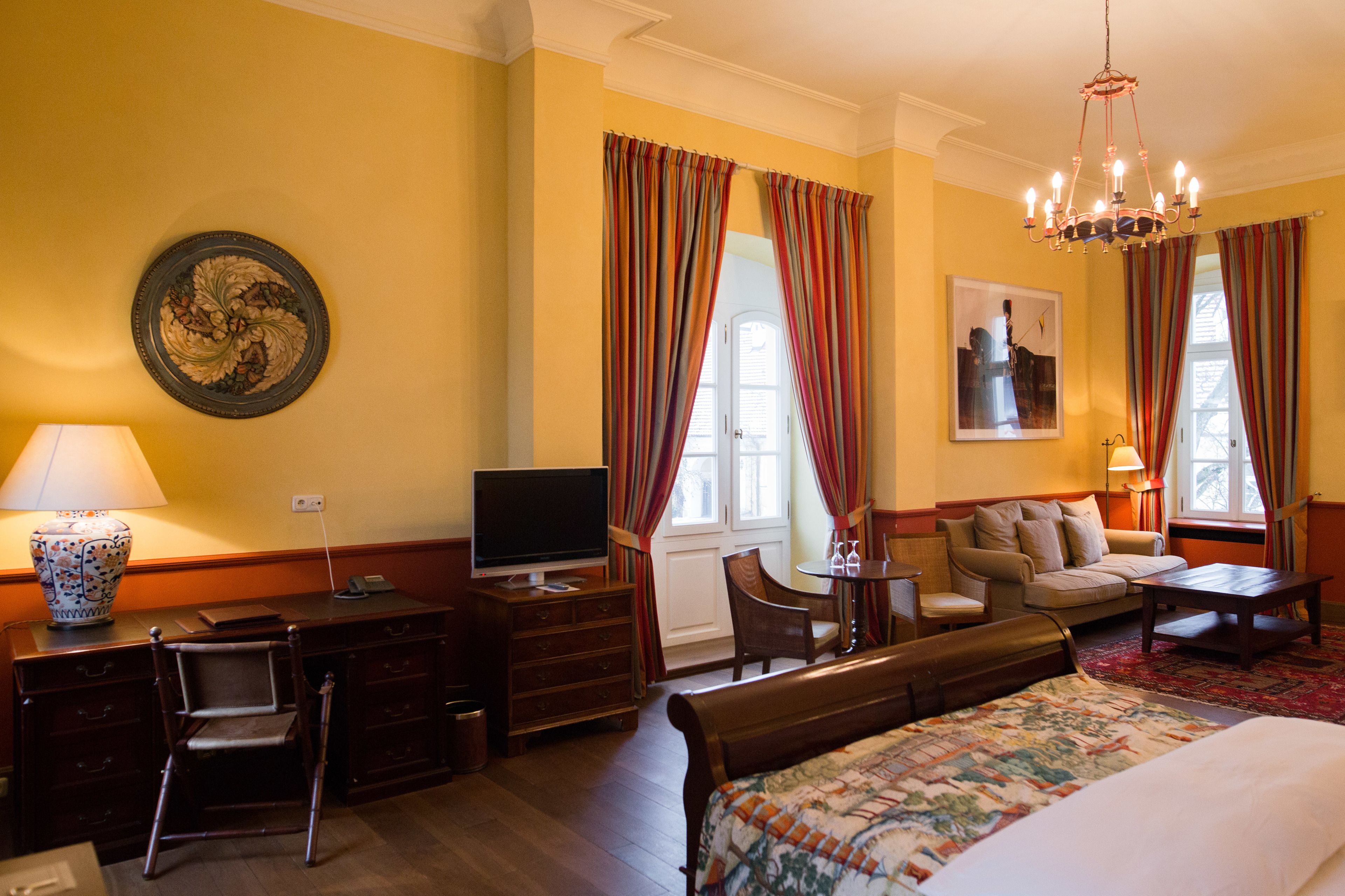 A hotel room with a bed, a desk with a television, a chair, a sofa, and a chandelier.
