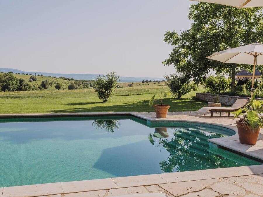 Una piscina con vistas a un campo verde y colinas. Hay plantas en macetas y una silla de descanso.