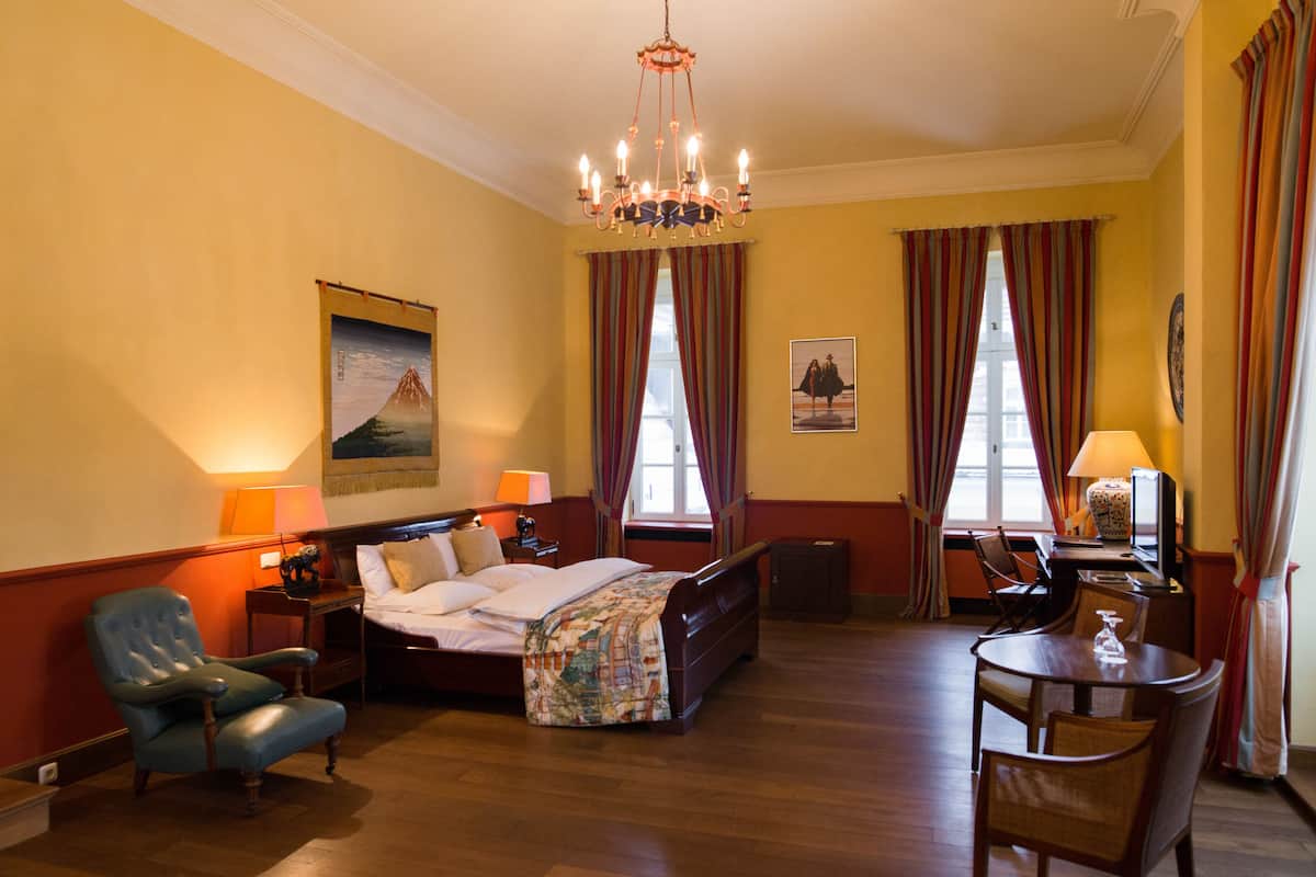 A hotel room with a bed, a desk, a chair, a lamp, and a chandelier.