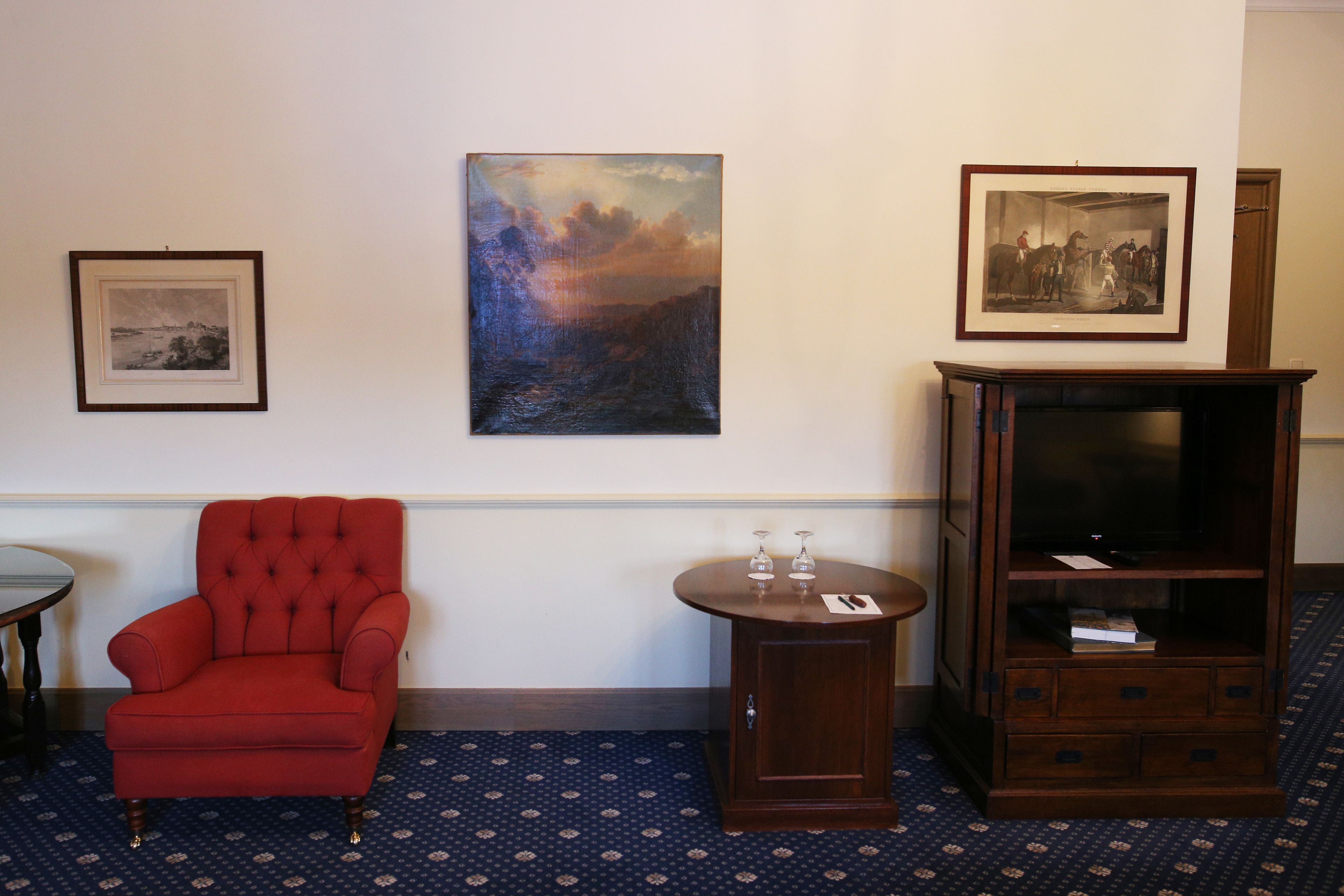 Una habitación con un sillón rojo, una mesita redonda con dos copas de vino, un mueble con televisor y dos cuadros en la pared.