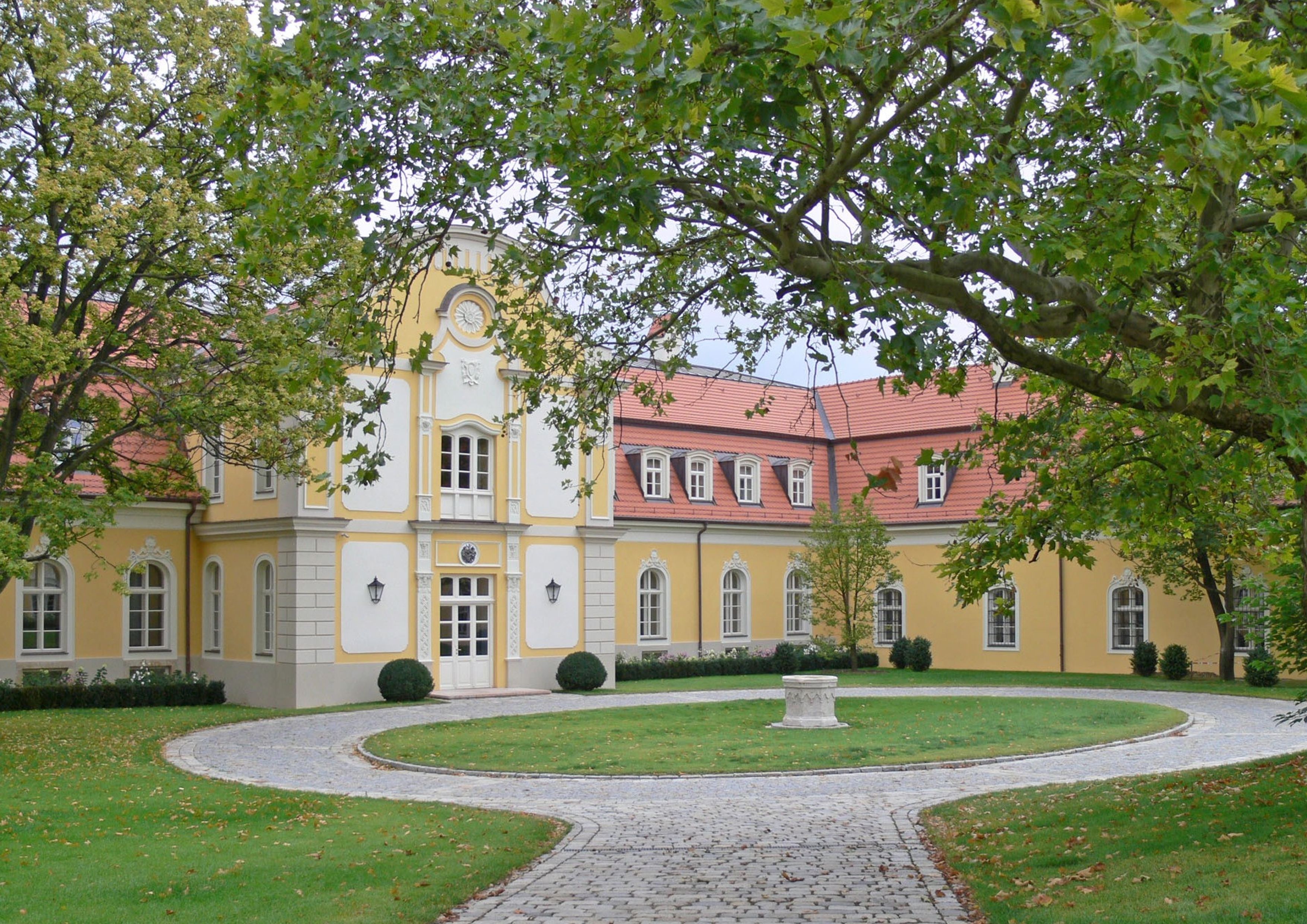 A historic building with a central clock tower, surrounded by a well-maintained garden with a paved pathway.