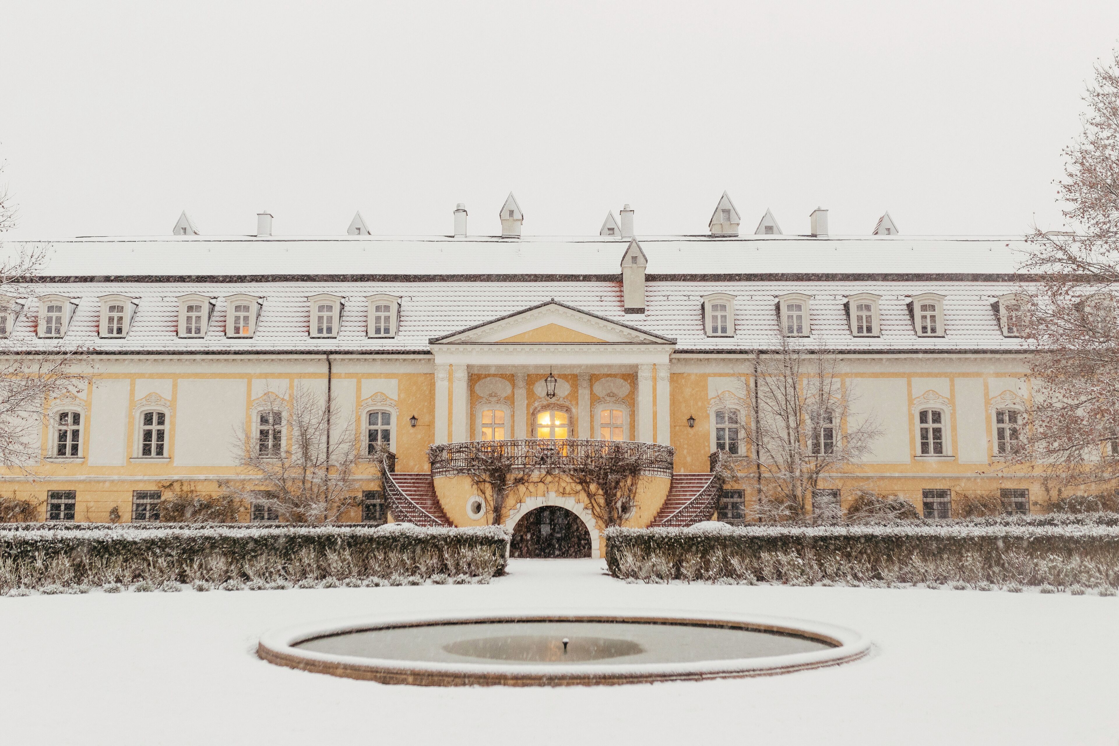 A snow-covered courtyard with a fountain, a building with a symmetrical facade, and a central entrance with a staircase.