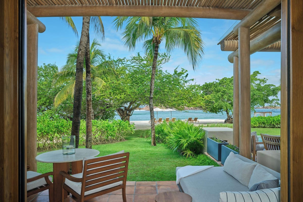 Un patio con una mesa redonda y sillas, un sofá, y vista a una playa y palmeras.