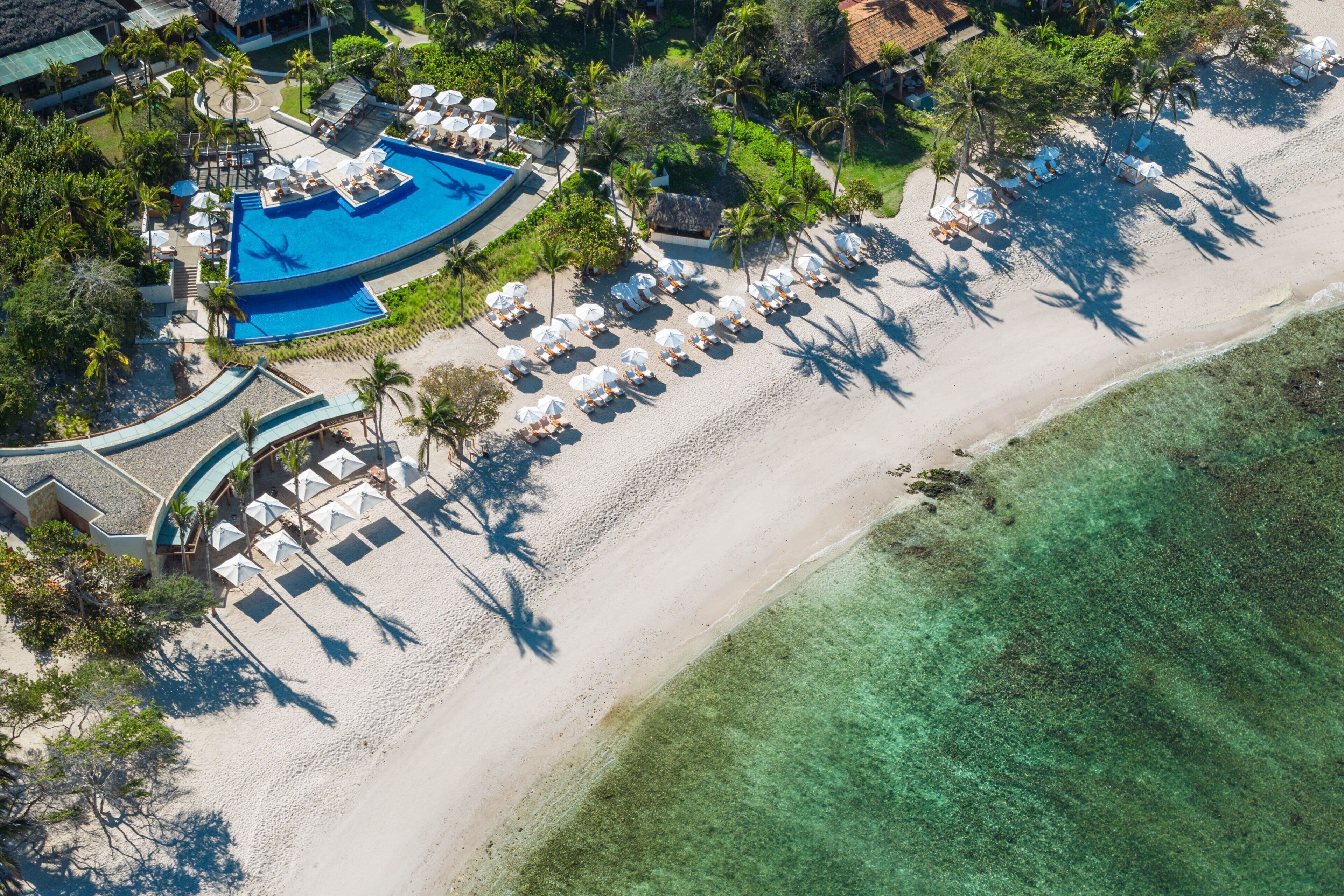 An aerial view of a resort with a swimming pool, beach, and lounge area with umbrellas.