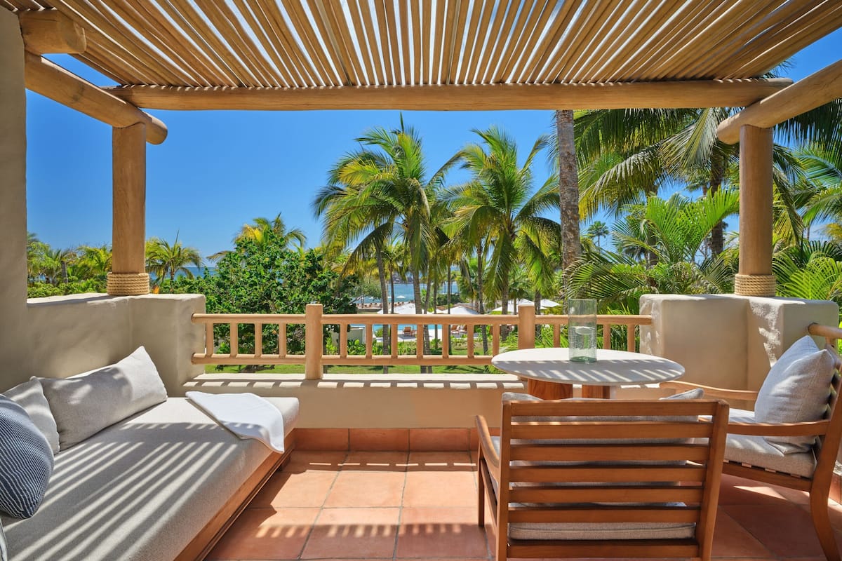 Un área de estar techada al aire libre, con vista a palmeras y una piscina.