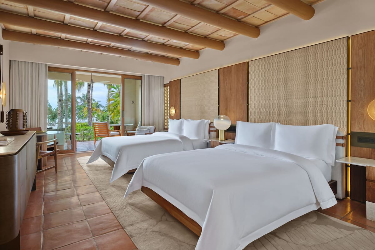 Habitación de hotel con dos camas, un escritorio y vistas a palmeras.