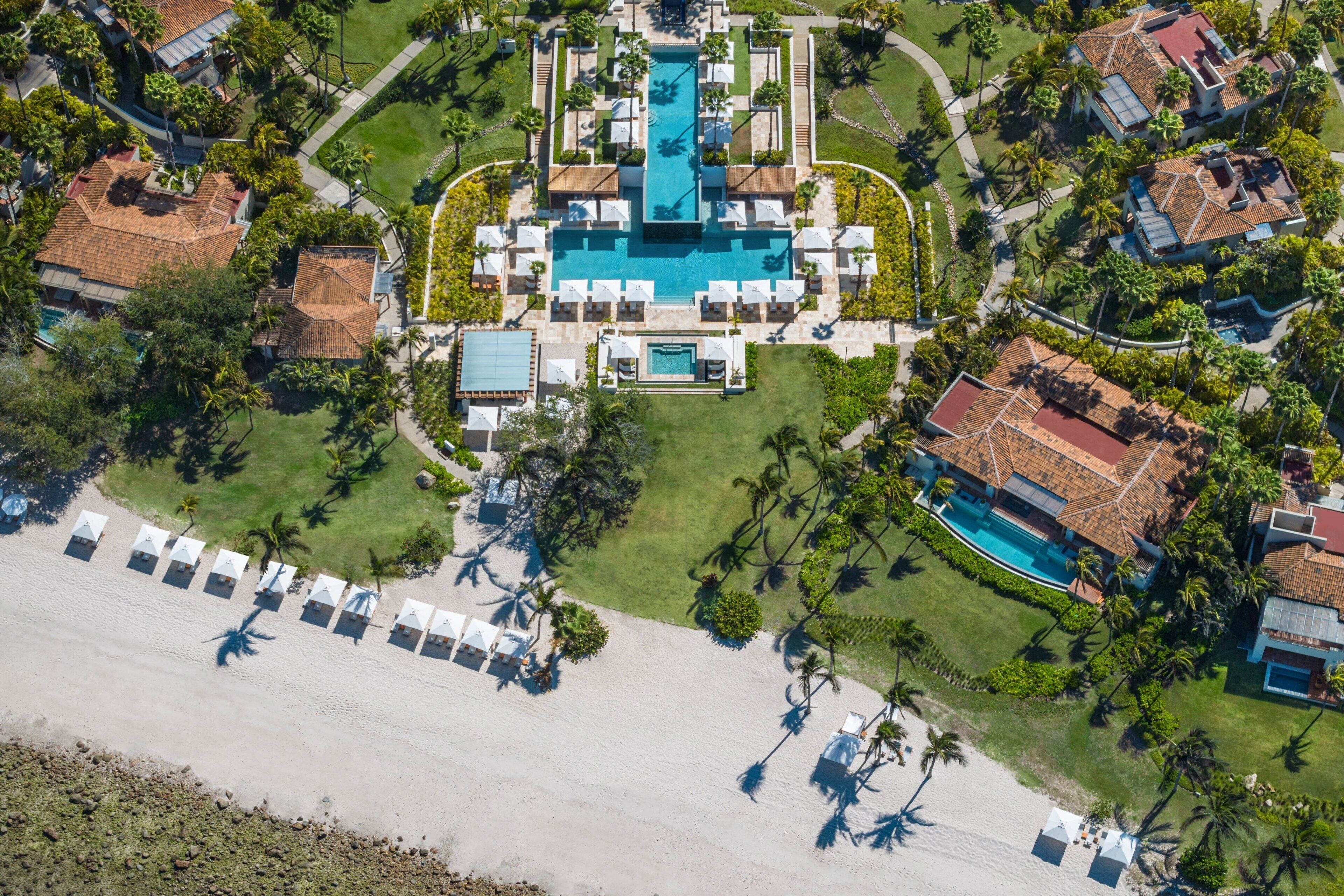 Vista aérea de un resort con piscina central, varias villas para huéspedes y una zona de playa.