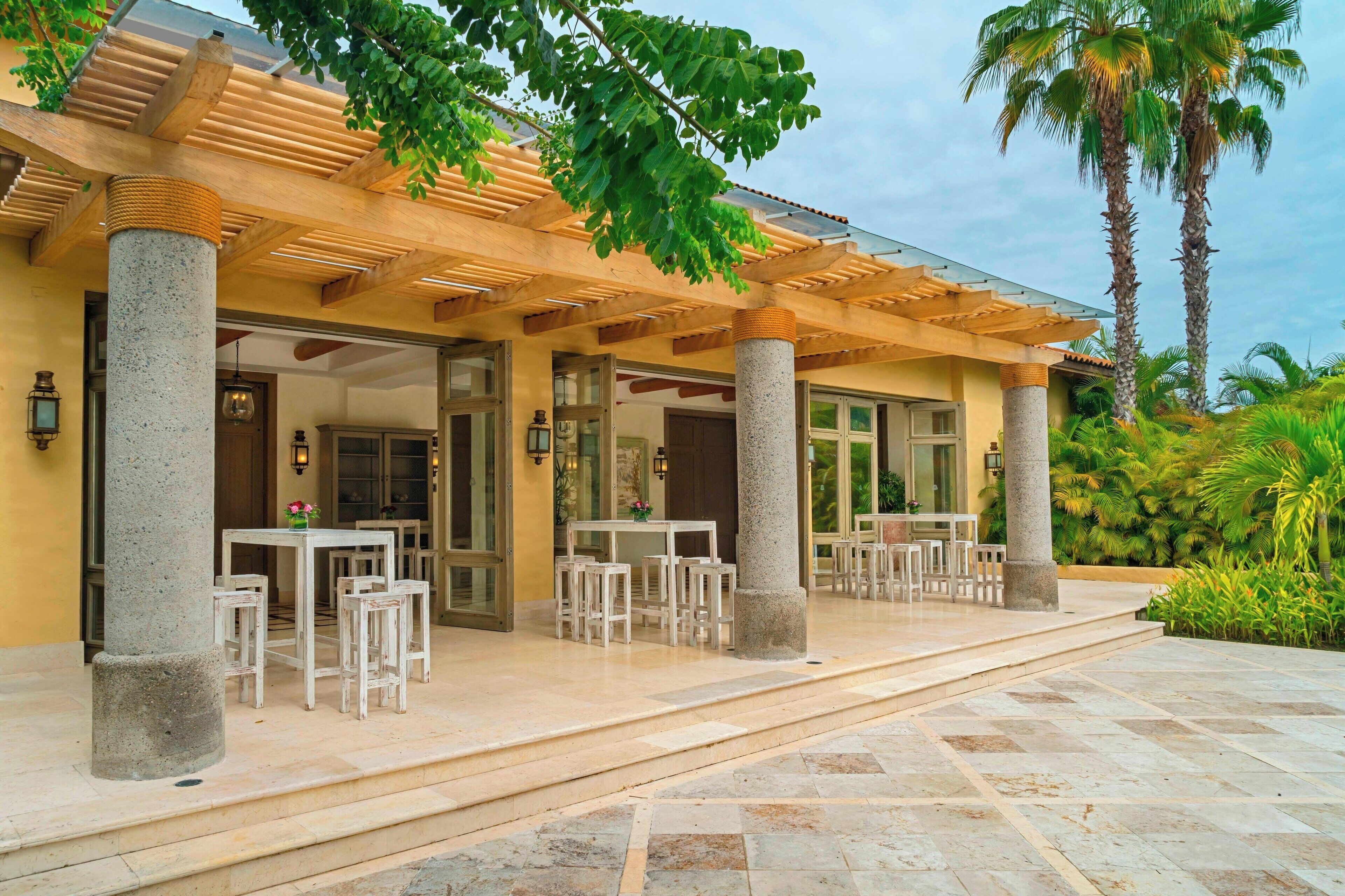 A covered outdoor seating area with stone columns, tables, and chairs, surrounded by palm trees and greenery.