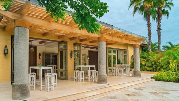 Un espace extérieur aménagé, abrité et doté de colonnes en pierre, de tables et de chaises, entouré de palmiers et de verdure.