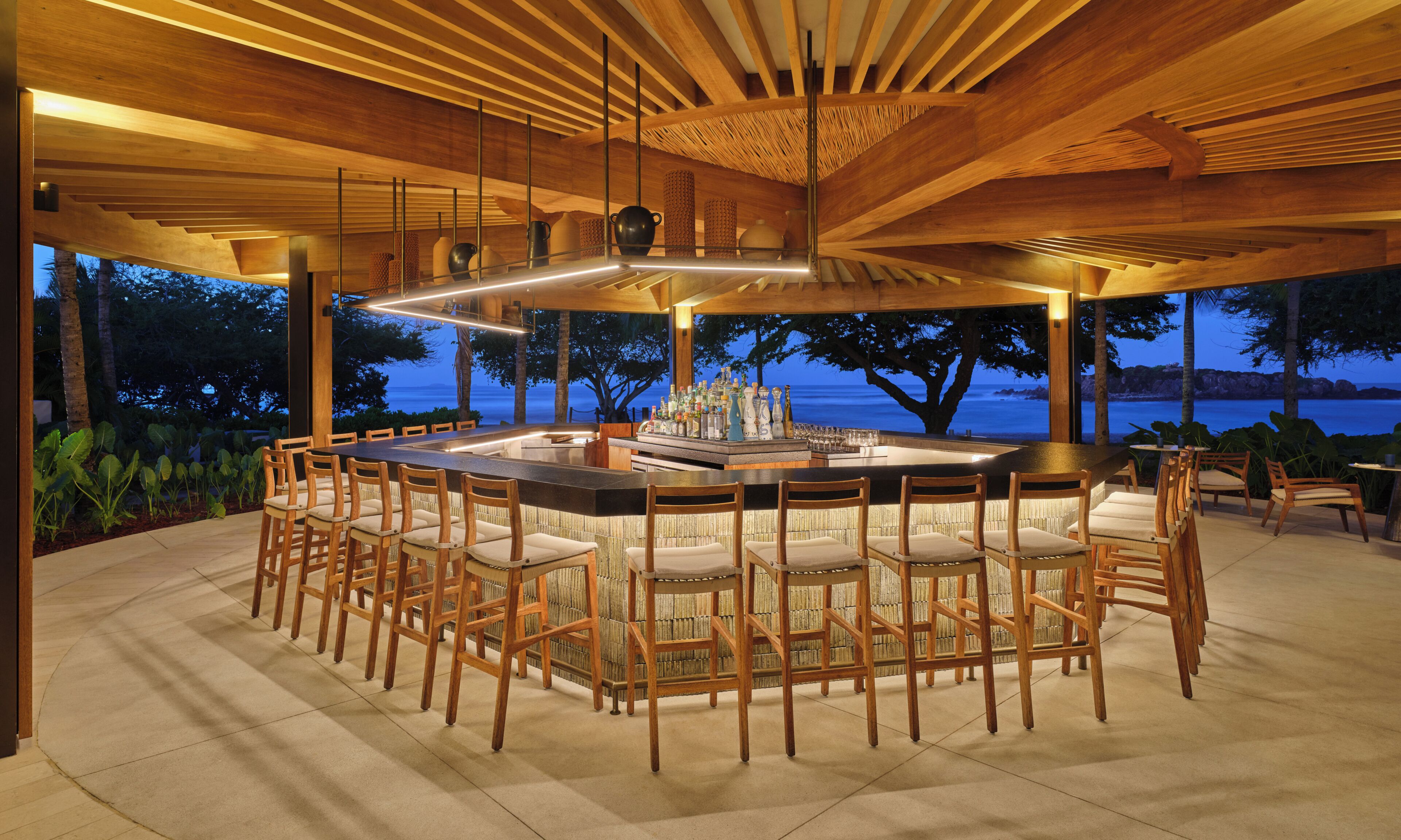 Una zona de bar con una mesa redonda y sillas, una barra con botellas y vistas al mar.