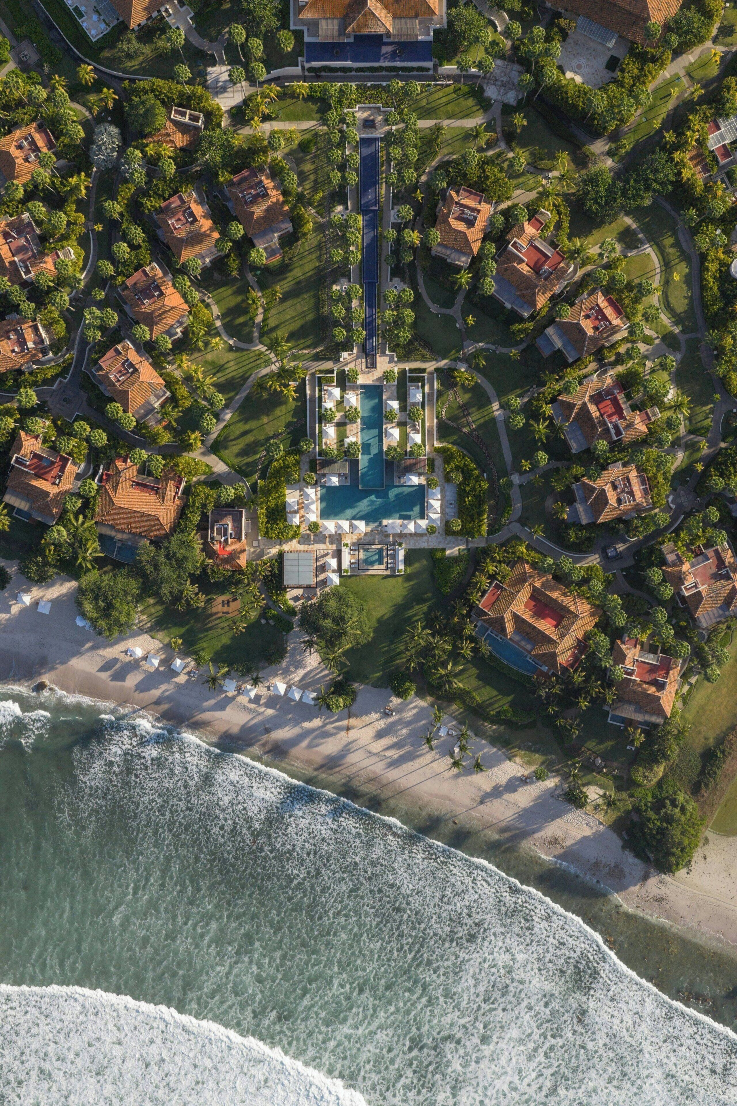 Vue aérienne d’un complexe hôtelier doté d’une piscine centrale, entouré de villas, d’une plage et bordé par une chaîne de montagnes.