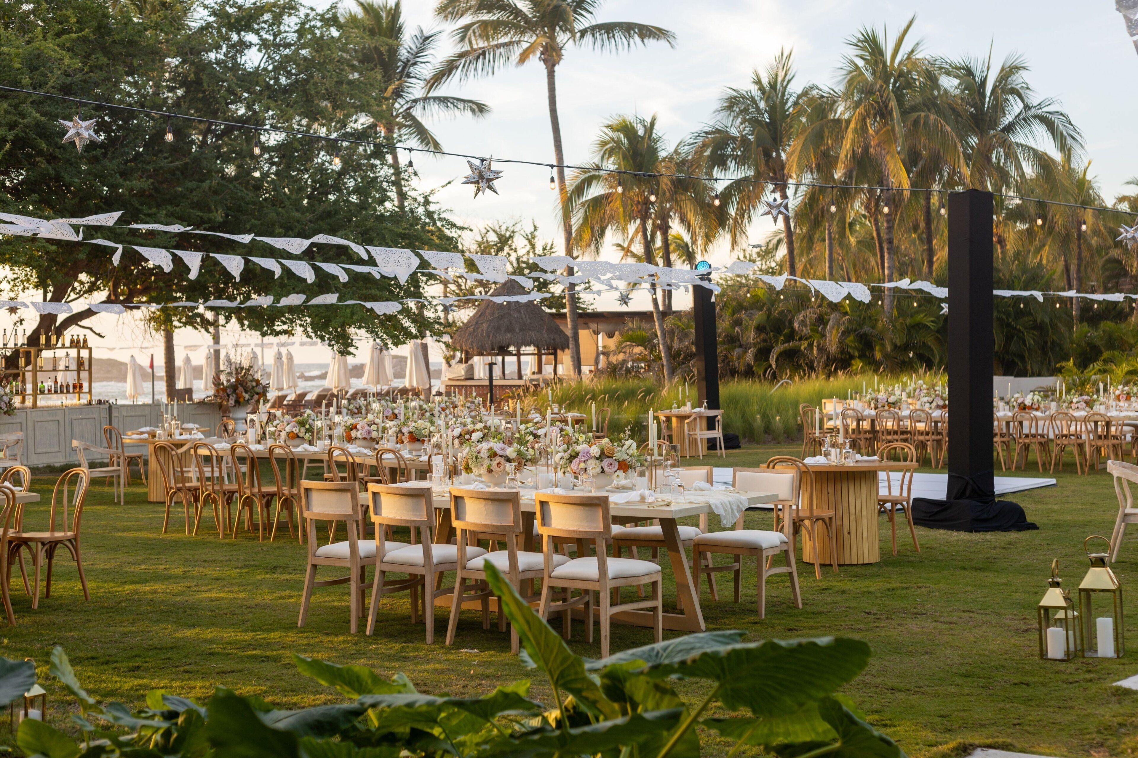 An outdoor event space with tables set for dining, surrounded by palm trees and a thatched-roof structure.