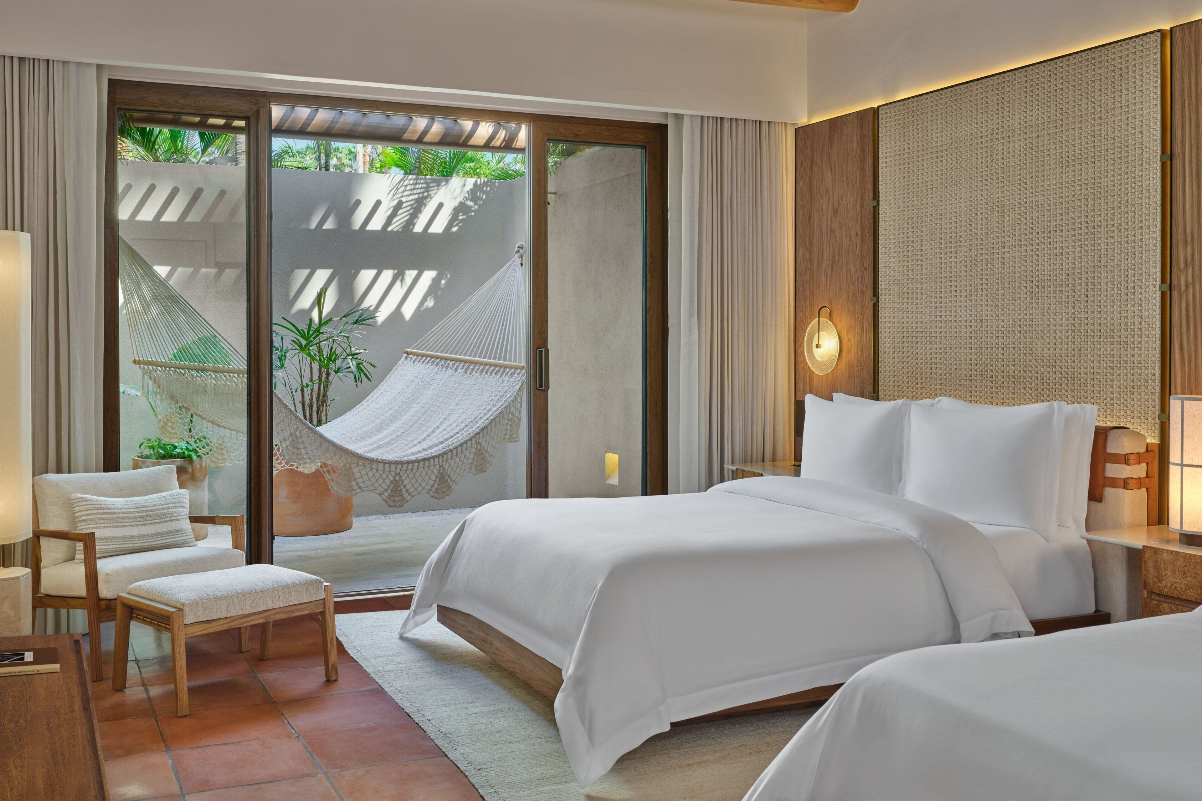 Une chambre d’hôtel moderne avec deux lits, un hamac, une chaise et une vue sur la verdure environnante.