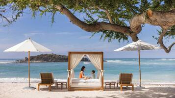 Escena de playa con una cama con dosel, dos sillas y una mesa bajo un árbol.
