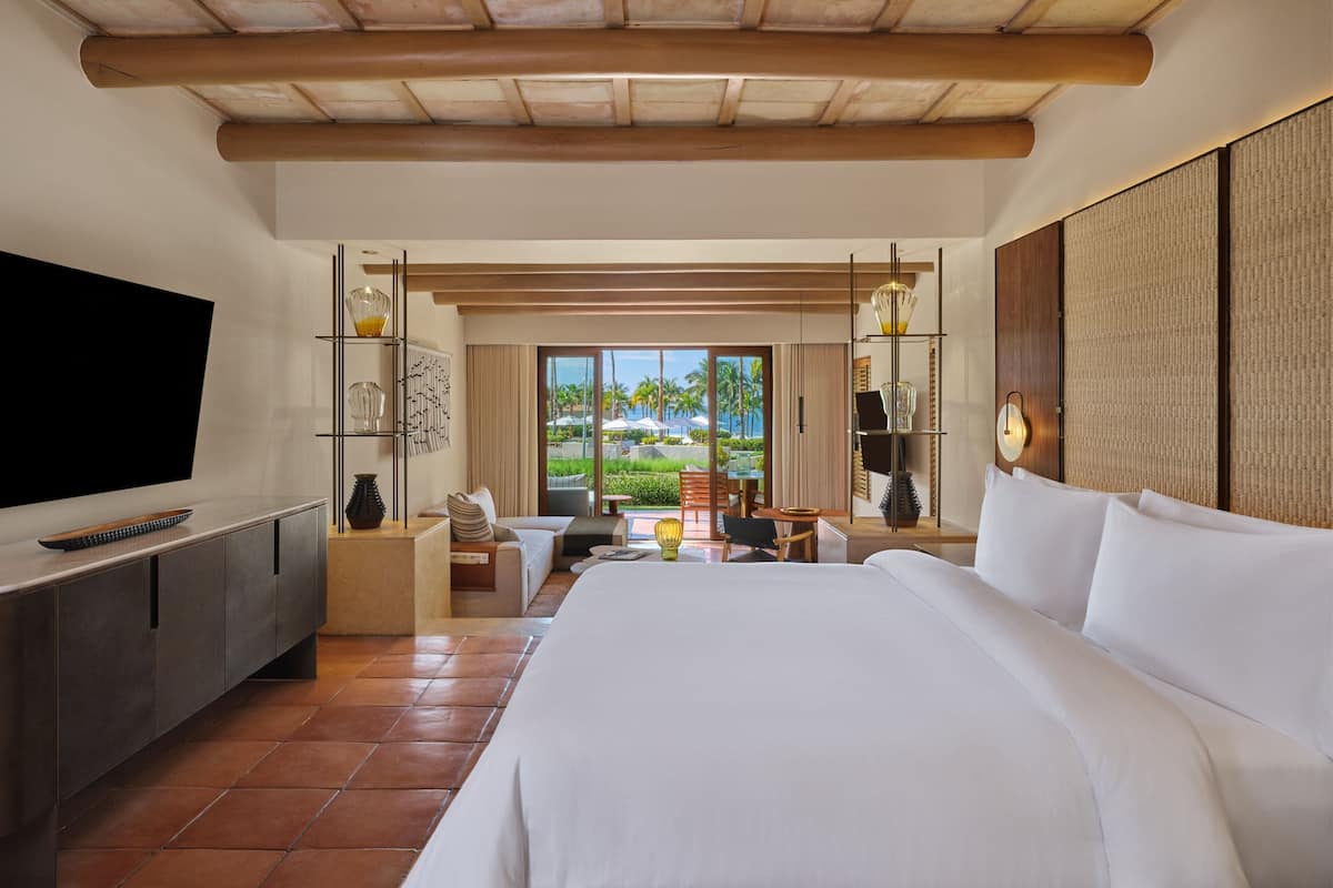 Habitación de hotel con una cama grande, televisión, escritorio con silla y vista a palmeras desde la ventana.
