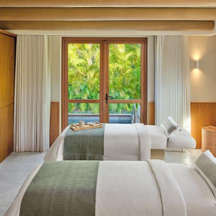 A hotel room with two beds, a large window with a view of greenery, a wooden bedside table with a plant, and a small side table with a lamp.