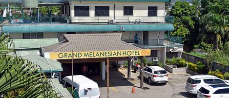 Hotel Grand Melanesian, un edificio de dos pisos, con estacionamiento techado y vistas a montañas al fondo.