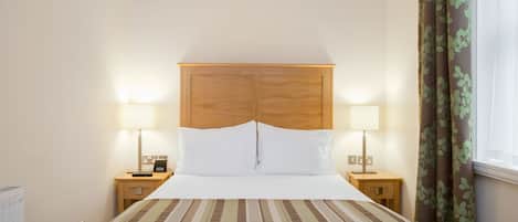 Une chambre d’hôtel avec un grand lit, deux tables de chevet avec des lampes, une tête de lit en bois et une fenêtre avec des rideaux.