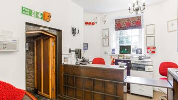 Una zona de recepción con un mostrador de madera, sillas rojas, un letrero de salida de incendios y una ventana con cortina floral.