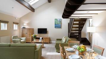 A spacious living room with a high ceiling, a flat-screen TV, a dining area with a table set for four, and a staircase leading to an upper floor.