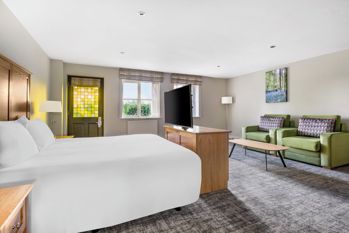 A hotel room with a bed, a TV, a desk, a chair, and a painting on the wall.