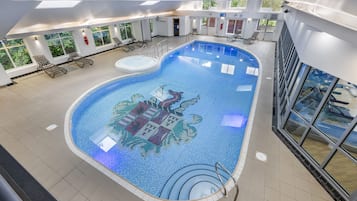 An indoor swimming pool with a mosaic design, sunlit windows, and lounge chairs.