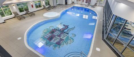 An indoor swimming pool with a mosaic design, sunlit windows, and lounge chairs.