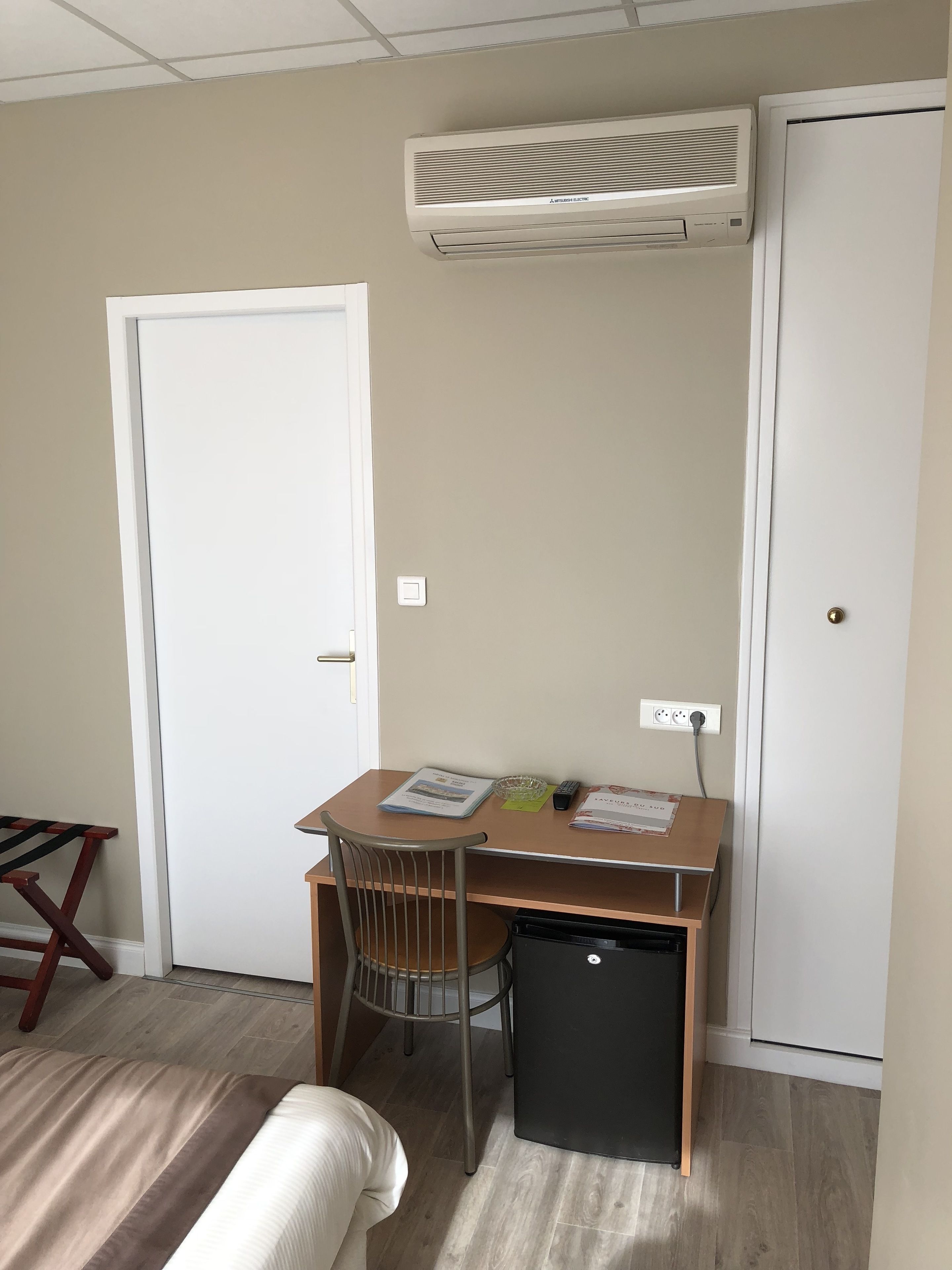 Een kleine kamer met een bureau, stoel en een compacte koelkast. Er is een witte deur en een airconditioner aan de muur bevestigd.