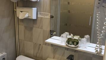 Baño con inodoro, lavabo y ducha. Hay toallas apiladas en un tendedero, un secador de manos y un dispensador de jabón.