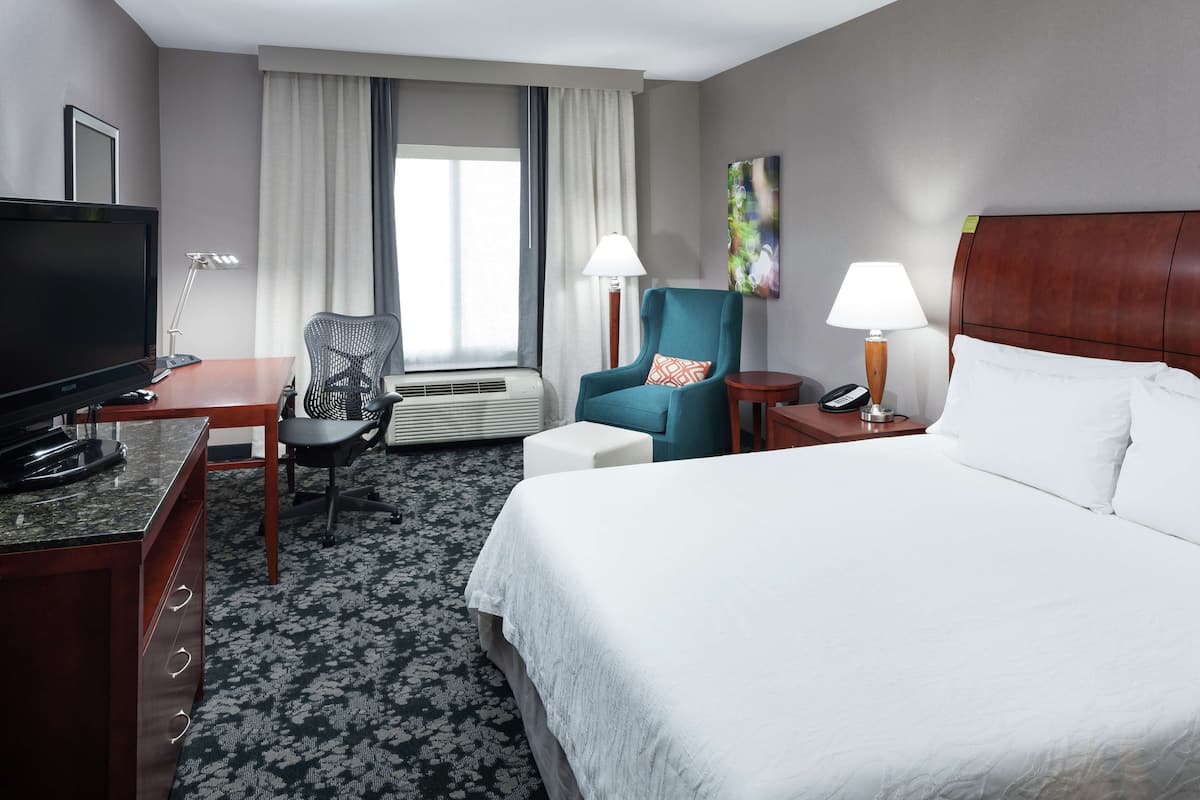 A hotel room with a bed, a desk with a TV, a chair, a lamp, and a window with curtains.