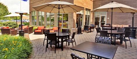 An outdoor patio area with brick flooring, outdoor seating with cushions, and umbrellas providing shade.