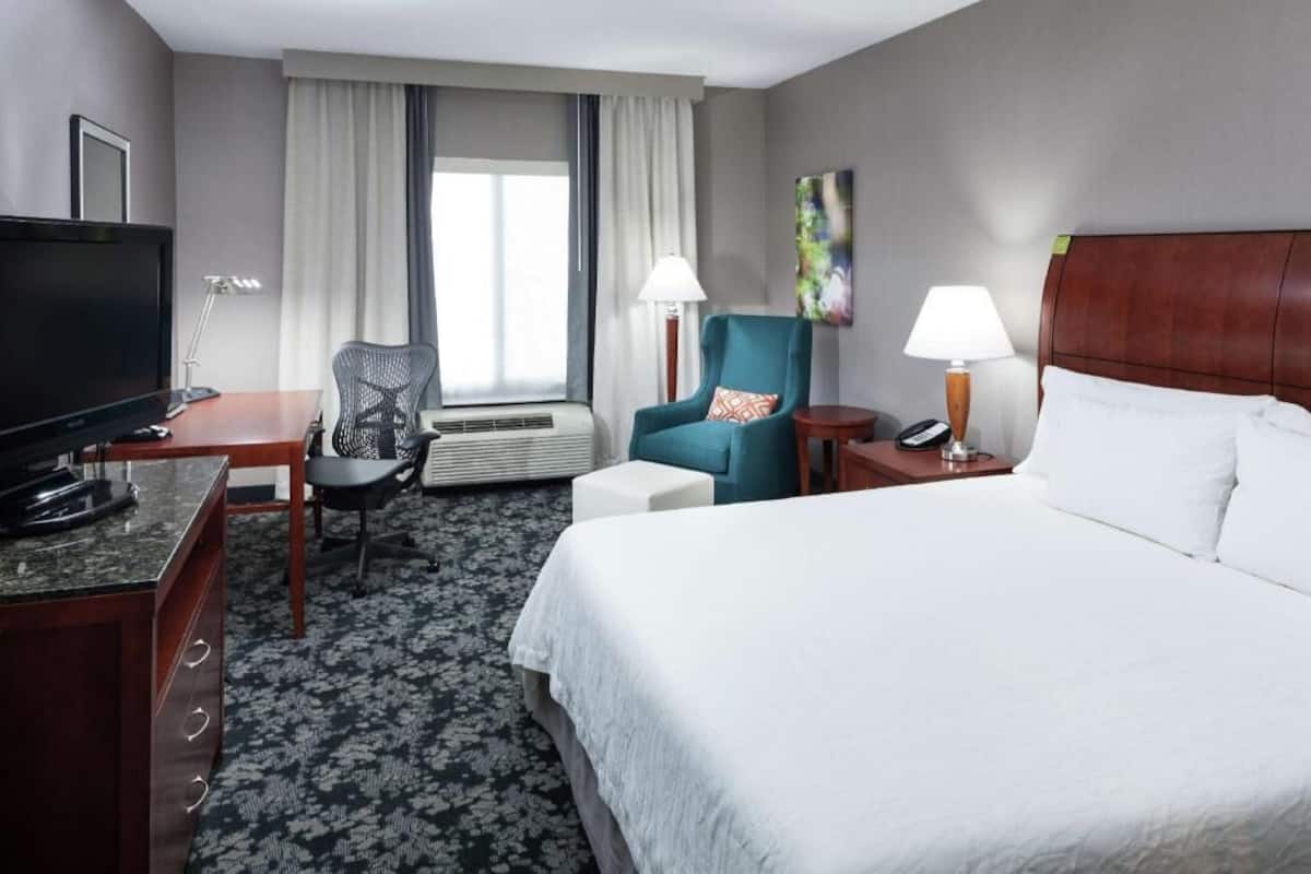 A hotel room with a bed, a desk with a TV, a chair, a lamp, and a window with curtains.