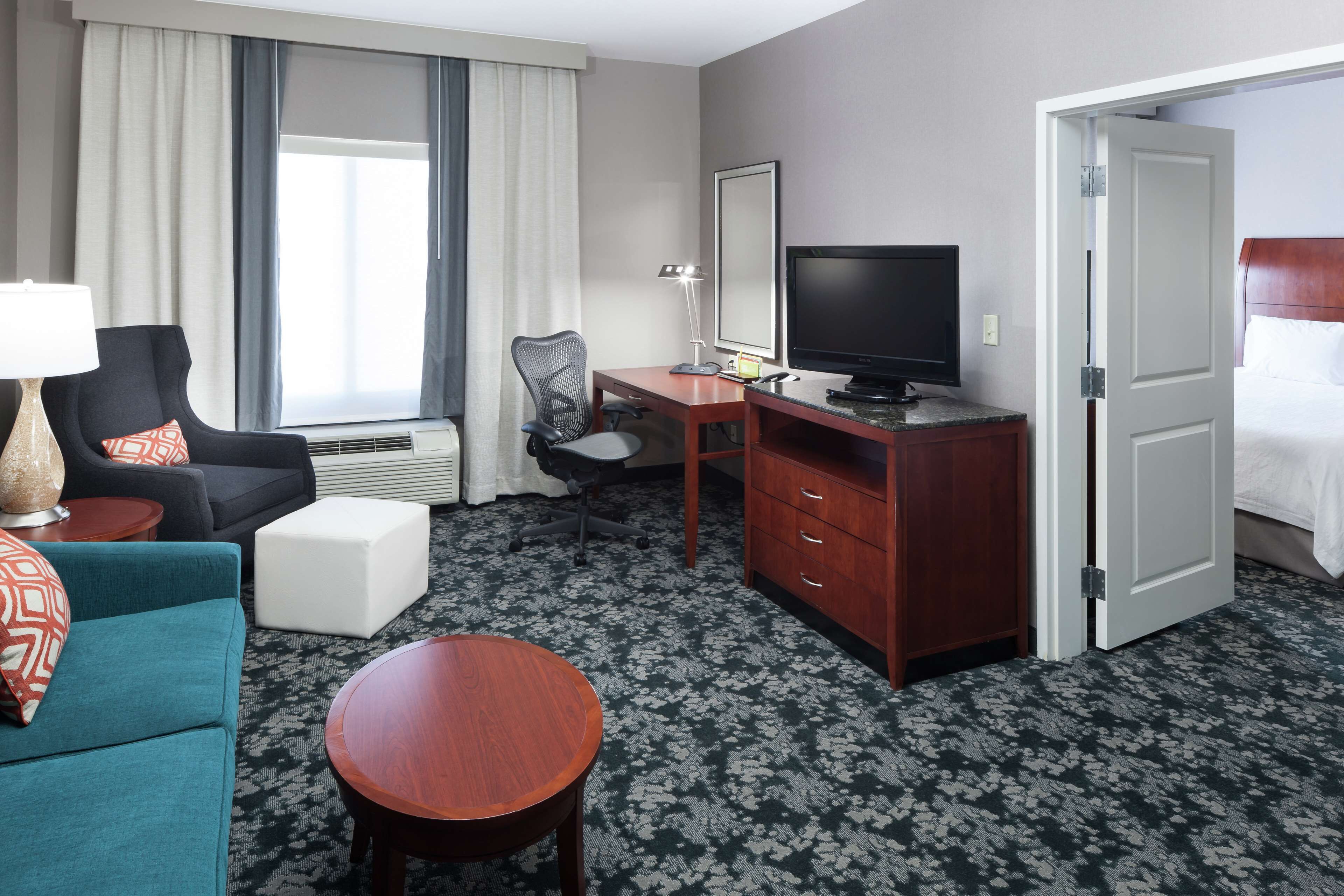 Ein Hotelzimmer mit Sofa, Schreibtisch mit Computer, Fernseher, Bett und einer Tür zu einem weiteren Raum.