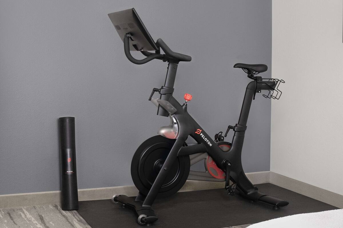 A stationary bike with a digital display and a red knob, placed in a corner with a grey wall and a black mat.