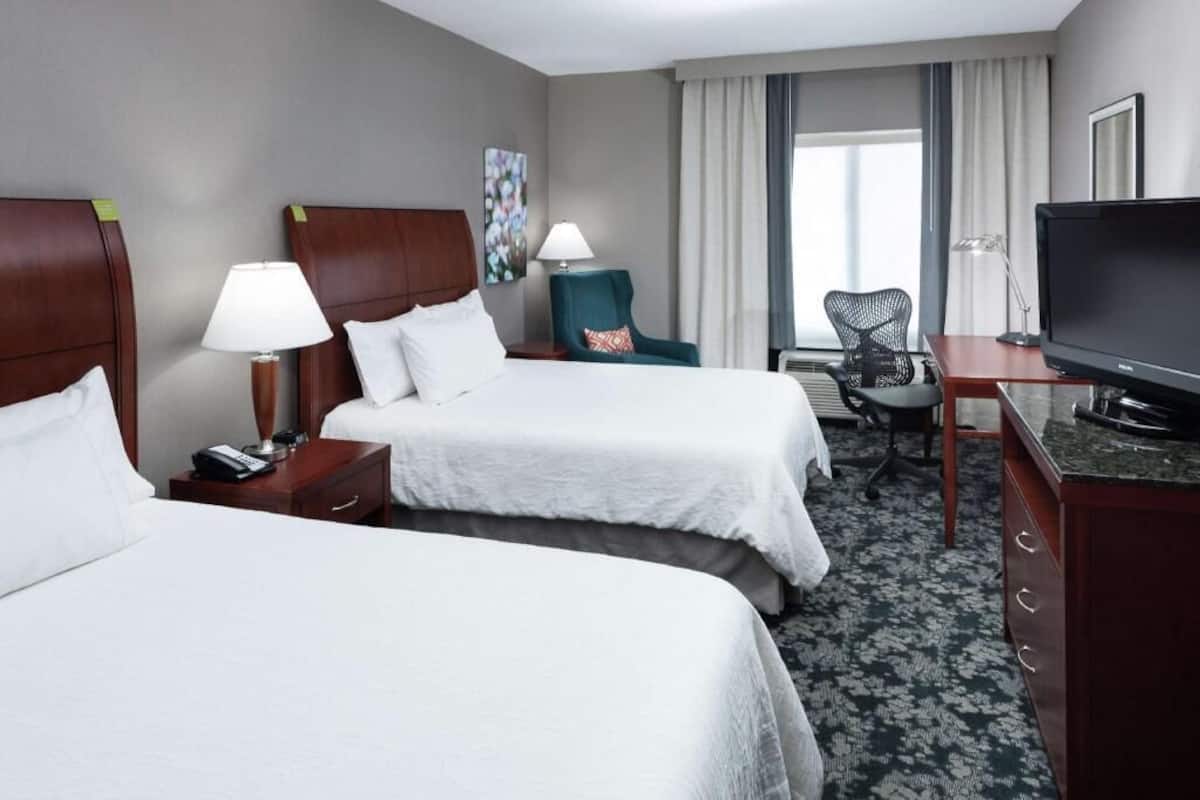 A hotel room with two beds, a desk with a TV, a chair, a lamp, and a painting on the wall.