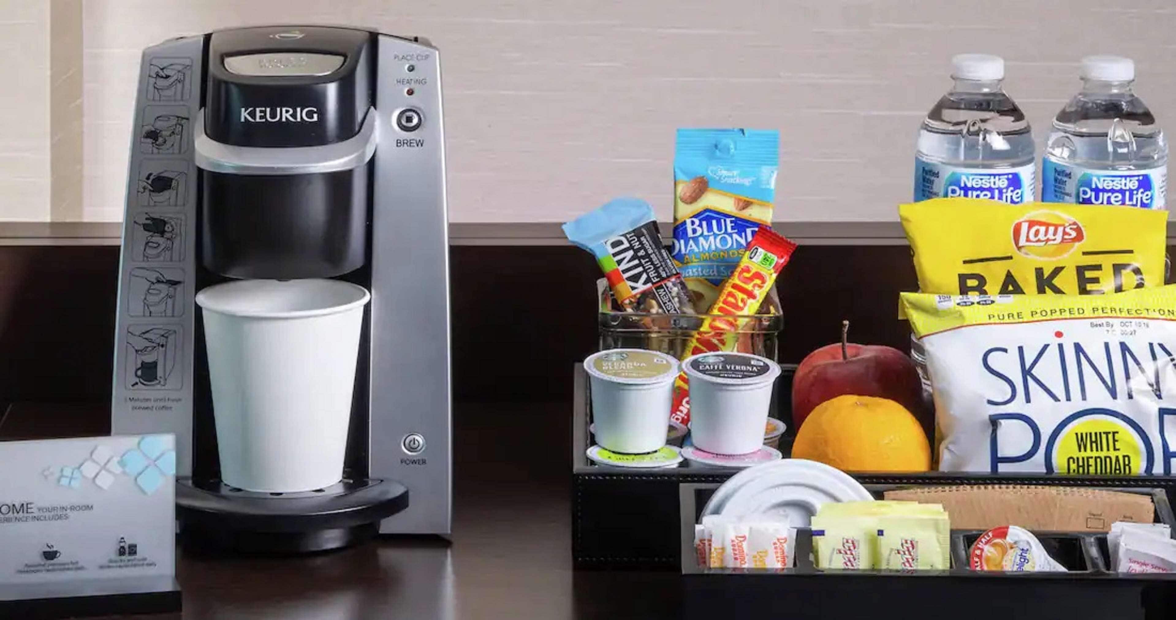 En Keurig-kaffemaskin med en kopp, ett fat med mat, inklusive snacks, och vattenflaskor.