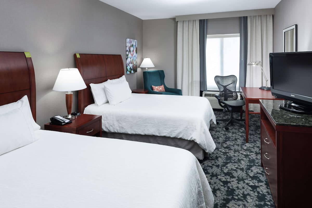 A hotel room with two beds, a desk with a television, a chair, and a lamp.