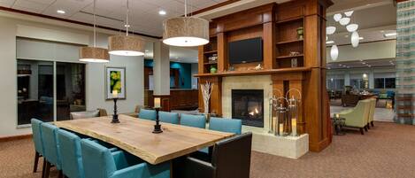 A spacious dining area with a large wooden table and blue upholstered chairs, a fireplace with decorative items, and a television mounted on a wooden wall.